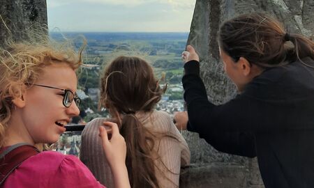 Eine Lehrerin und zwei Schülerinnen stehen an einer Maueröffnung. Die Lehrerin zeigt einer Schülerin etwas unten im Tal.