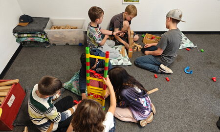 Sechs Kinder bauen und konstruieren Türme aus verschiedenen Materialien, wie bunten Röhren und Holzklötzen. Sie sitzen auf einem Teppichboden.