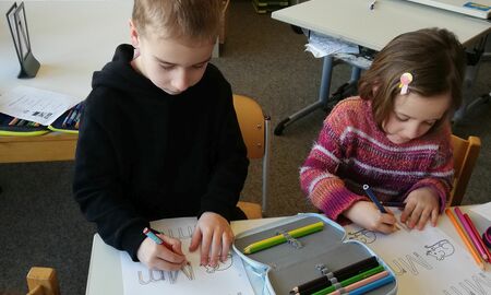 Ein Junge und ein Mädchen sitzen gemeinsam an einem Tisch. Sie schreiben mit Buntstiften den Groß- und Kleinbuchstaben M bunt.