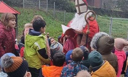Der Nikolaus steht mit einem roten Buch umringt von einer Gruppe Erstklässlerinnen und Erstklässlern auf einem Spielplatz.