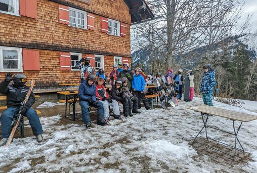 Schüler sitzen auf Bierbänken vor dem historischen Schöntalhof. Sie sind für das Snowboarden und Skifahren eingekleidet. Es liegt Schnee.