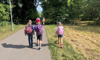Drei Kinder sind vorne im Bild beim Wandern auf einem Feldweg zu sehen. Im Hintergrund sieht man noch mehr Kinder auf dem Feldweg.