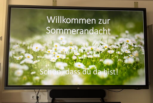 Auf einer digitalen Tafel steht der Schriftzug "Willkommen zur Sommerandacht. Schön, dass du da bist!" Im Hintergrund des Schriftzuges ist eine Wiese mit Gänseblümchen zu sehen.