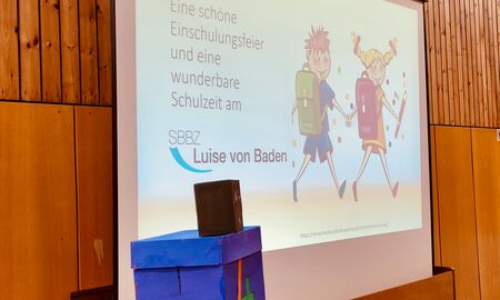 Präsentation auf einer Leinwand mit dem Text "Eine schöne Einschulungsfeier und eine wunderbare Schulzeit am SBBZ Läuse von Baden" und zwei Cliparts von Schulkindern, die einen Jungen und ein Mädchen mit Schulranzen darstellen.