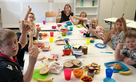 Eine Lehrerin sitzt mit sieben Kindern der zweiten Klasse im Klassenzimmer an einem langen und reich gedeckten Frühstückstisch. Alle zeigen einen Daumen hoch.