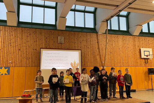 Einige Kinder der zweiten Klasse stehen auf der Bühne, tragen Fürbitten vor und singen dazu ein Lied, von welchem im Hintergrund der Text über den Beamer zu sehen ist.