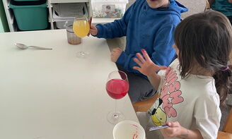 Zwei Kinder sitzen an einem Tisch, vor ihnen stehen zwei Gläser mit einer bunten Flüssigkeit drin, die Kinder schauen auf die Finger von dem Kind, das weiter vorne im Bild sitzt, die Finger sind rot gefärbt, im Hintergrund ist ein Klassenzimmer zu sehen