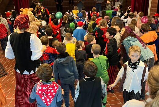 Auf der Bühne der Aula tanzen Lehramtsanwärterinnen vor und davor stehen die verkleideten Schülerinnen und Schüler der Grundschule Sprache.