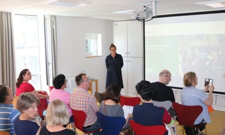 Ein Publikum schaut bei einem Vortrag auf eine Leinwand. Die Referentin steht vorne
