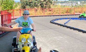 Ein Junge fährt mit einem Go-Kart auf einer Kartbahn. Links im Bild werden Hände ausgestreckt. Der Junge auf dem Go-Kart hält ebenfalls seine Hand raus, um einzuschlagen