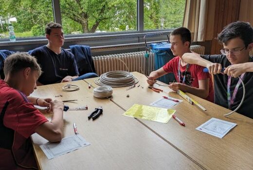 Vier Jungs sitzen um einen Tisch mit einer Rolle Kabel Zangen und einem Leitungsprüfer. Einer isoliert gerade ein Kabel ab.