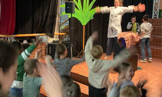 Eine Lehrerin springt rechts im Bild mit beiden Armen ausgestreckt in die Luft auf einer Bühne, links im Bild stehen viele Kinder vor ihr und springen ebenfalls in die Luft