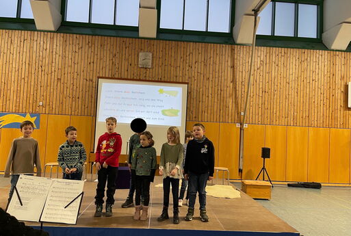 Acht Kinder der zweiten Klasse singen Stern über Betlehem, stehen dabei auf einer Bühne und im Hintergrund ist über den Beamer der Text zu sehen.