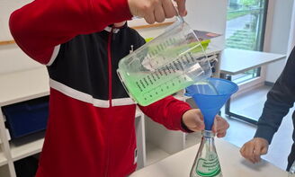 Ein Junge steht an einem Tisch und schüttet mit einem Messbecher eine grüne Flüssigkeit mithilfe eines Trichters in eine Glasflasche