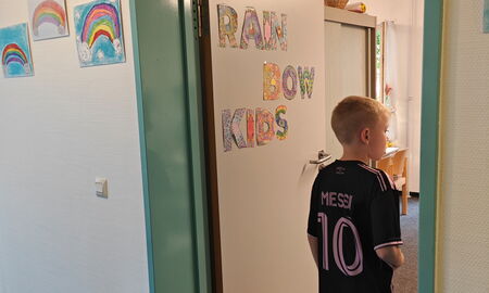 Eine Tür mit der Aufschrift "Rainbow Kids" steht offen, ein Junge geht grade hinein. Links neben der Tür sind drei Bilder von Regenbögen