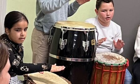 Eine Schülerin und zwei Schüler sitzen oder stehen an unterschiedlichen Trommeln und trommeln einen Rhythmus.