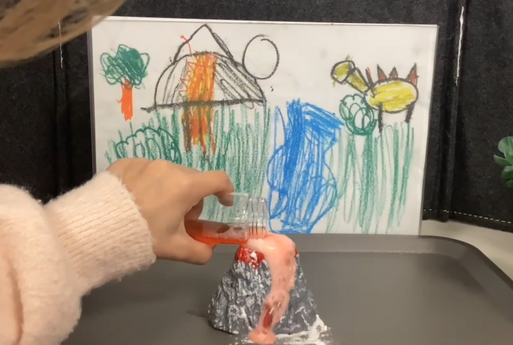 In der Mitte des Bildes sieht man einen Mini-Vulkan, ein Kind schüttet Flüssigkeit hinein, aus dem Vulkan kommt eine schaumige Flüssigkeit raus, im Hintergrund sieht man ein selbst gemaltes Bild