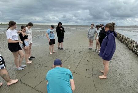 Zehn Personen, LehrerInnen und SchülerInnen, stehen um ein in den Strand gezogenes Rasterfeld. In der Einzelfeldern liegen Fundstücke aus dem Watt.