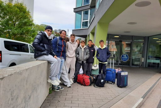 Die Mannschaft posiert zu Anfang vor einem Gebäude. Vor und neben der Gruppe stehen ihre Koffer und Taschen.