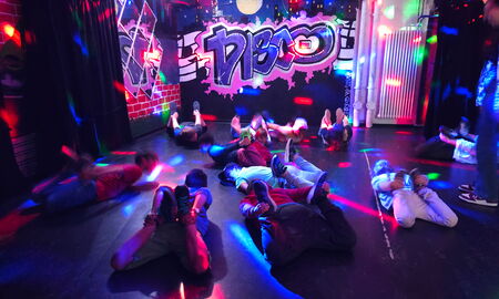 Einige Kinder liegen in einem Raum mit dem Schriftzug "Disco" bei buntem Discolicht auf dem Bauch und halten ihre Füße fest.