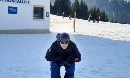 Ein Junge steht im Schatten vor dem Lifthaus auf dem Snowboard, er ist in die Knie gegangen und lacht