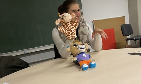 Eine Lehrerin hält Leo, eine Löwenhandpuppe und eine Giraffenhandpuppe in die Höhe. Sie sitzt an einem Tisch.