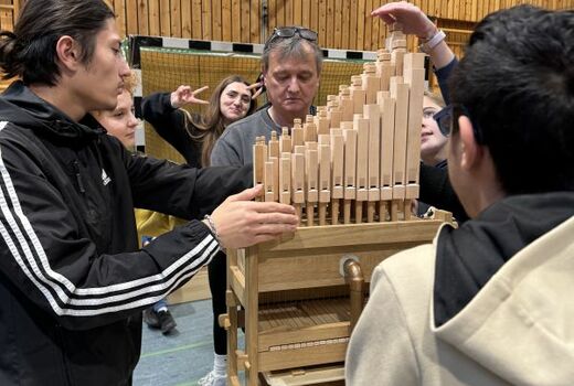 Ein Mann spielt die fertige Orgel. Ein Junge betätigt die Blasebälge und einer hält eine Hand vor die Orgelpfeifen, um die Töne zu erspüren.