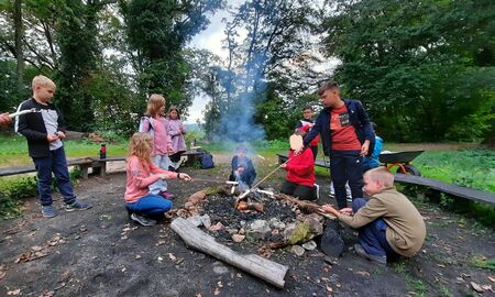 Neun Kinder und eine Lehrkraft stehen und sitzen um ein Lagerfeuer herum. Vier Kinder halten Stöcke mit Stockbrot über das kleine Feuer.