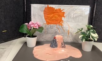 In der Mitte des Bildes sieht man einen Mini-Vulkan auf einem Tablett stehen, aus dem Vulkan kommt eine schaumige Flüssigkeit raus, im Hintergrund sieht man ein selbst gemaltes Bild von einem Vulkan, links und rechts stehen Blumen