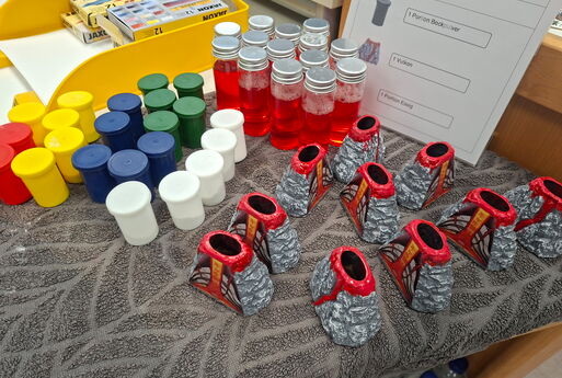 Auf einem Tisch stehen kleine Vulkane, verschiedenfarbige Filmdosen und Behälter mit einer roten Flüssigkeit