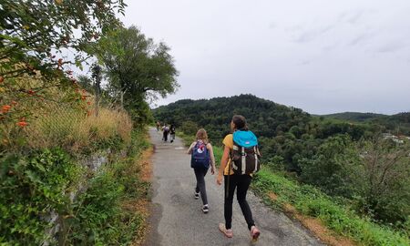 Eine Lehrerin und Kinder laufen auf einem asphaltierten Weg. Sie tragen Rucksäcke. Rechts und links sind Wiesen und Bäume.