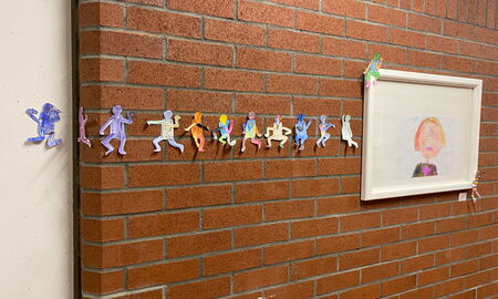 11 bunte kleine Papiermännchen hängen in einer langen Reihe an der Wand. Recht daneben hängt ein Bild, an dem auch zwei Papiermännchen kleben.