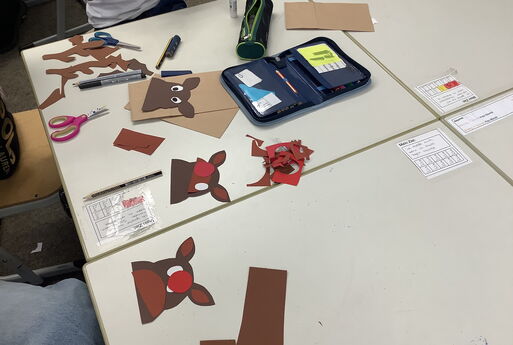 Auf einem Tisch in der Klasse liegen verschiedene Teile aus Tonkarton für das Adventskalender-Tütchen, die einen Rentier-Kopf bilden sowie Schere, Kleber und Mäppchen