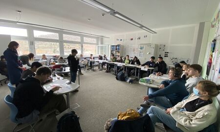 Eine Schulklasse sitzt im Halbrund. Vorne, zu ihnen gewandt, steht ein Schüler und spricht in ein Mikrofon.