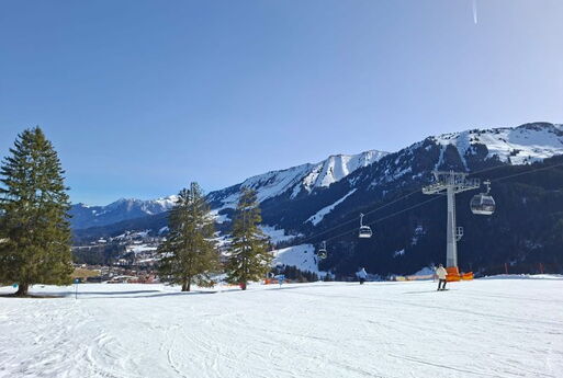 Ein verschneiter Skihang in den Bergen. Rechts ein Lift und ein paar Skifahrer.