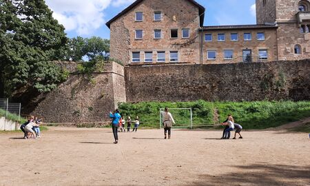 Auf einer sandigen Fläche vor einem alten Gebäude machen zwei Kindergruppen von drei bis vier Kindern tauziehen. Eine dritte Schülergruppe schaut zu. Eine Lehrerin feuert an, die andere zeigt die Mitte an.