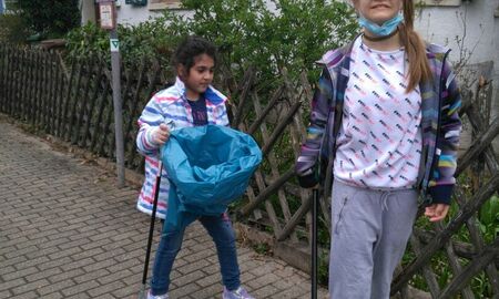 Zwei Mädchen sammeln Müll auf der Schützenhausstraße ein. Beide haben eine Zange in der Hand. Ein Mädchen zusätzlich eine blaue Tüte. Sie stehen auf dem Gehweg.