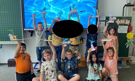 Die Kinder der Drachenklasse stehen vor einer interaktiven Tafel mit Wasserbild, halten ihre Wasserfahrzeuge hoch und posieren.