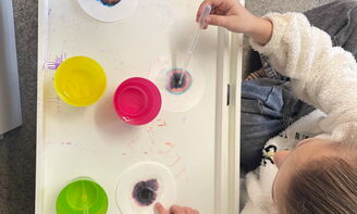 Aus der Vogelperspektive sieht man drei Kinder, die vor einem weißem Tablett sitzen, zwei Kinder tropfen mit einer Pipette Wasser auf ein zum Teil bunt gefärbtes Papier, ein Kind zeigt auf sein Papier, auf dem Tablett stehen außerdem drei Wasserbecher