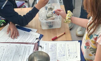 Drei Kinder stehen an einem Tisch und halten einen Gegenstand in einen Behälter, vor ihnen liegen beschriftete Blätter Papier auf dem Tisch, daneben steht ein Alubehälter mit einer Flüssigkeit