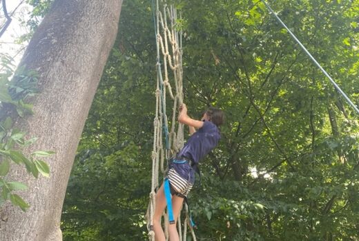 Ein Mädchen, gesichert durch einen Klettergurt und ein Seil, klettert an einem Netz nach oben.