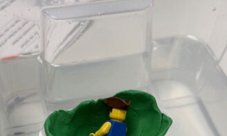 In einem Behälter mit einer Flüssigkeit schwimmt eine kleine grüne Schale, darin liegt eine Lego-Figur