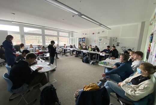 Eine Schulklasse sitzt im Halbrund. Vorne, zu ihnen gewandt, steht ein Schüler und spricht in ein Mikrofon.