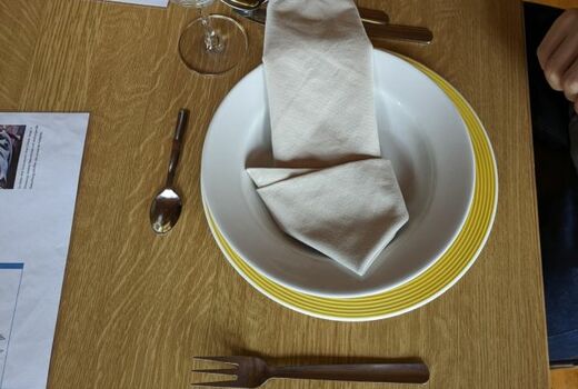 Ein Gedeck auf einem Tisch: Zwei Teller, eine Serviette, ein Glas, Messer, Gabel, Löffel, Dessertlöffel.