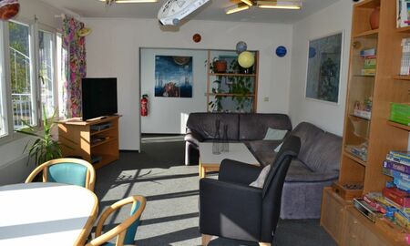 Man sieht das Wohnzimmer einer Gruppe mit Esstisch, Sofas und Sessel, Fernseher und Schrankwand mit vielen Brettspielen