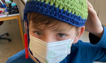 Ein Schüler mit Maske und einer selbstgehäkelten Mütze auf dem Kopf an der noch eine Nadel hängt