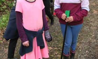 Zwei Drittklässlerinnen mit Handschuh und Müllzange schauen in die Kamera und im Hintergrund laufen weitere Drittklässler mit Handschuhen auf einem Waldweg und suchen nach Müll.
