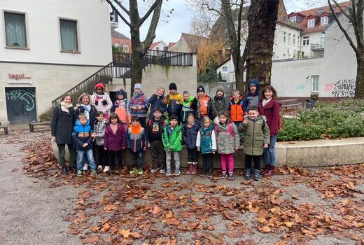 Zwanzig Schülerinnen und Schüler aus zwei ersten Klassen posieren mit zwei Lehrerinnen und einer Erzieherin vor einem Baum in der Stadt für ein Gruppenfoto.