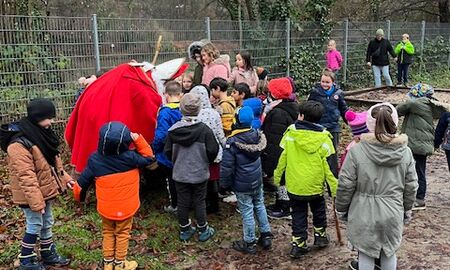 Der Nikolaus ist umringt von Erklässlerinnen und Erstklässlern auf einem Spielplatz und bückt sich, um die Geschenke hervorzuholen.