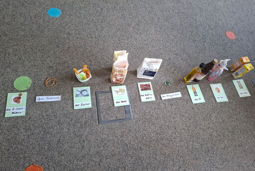 Auf dem Boden liegen in einer Reihe eine 2-Cent-Münze, ein Gummiband, Zucker, Mehl, Kaffeepulver, Muggelsteine, Öl, Seife und Salz, davor liegen Karten, die die Gegenstände beschriften